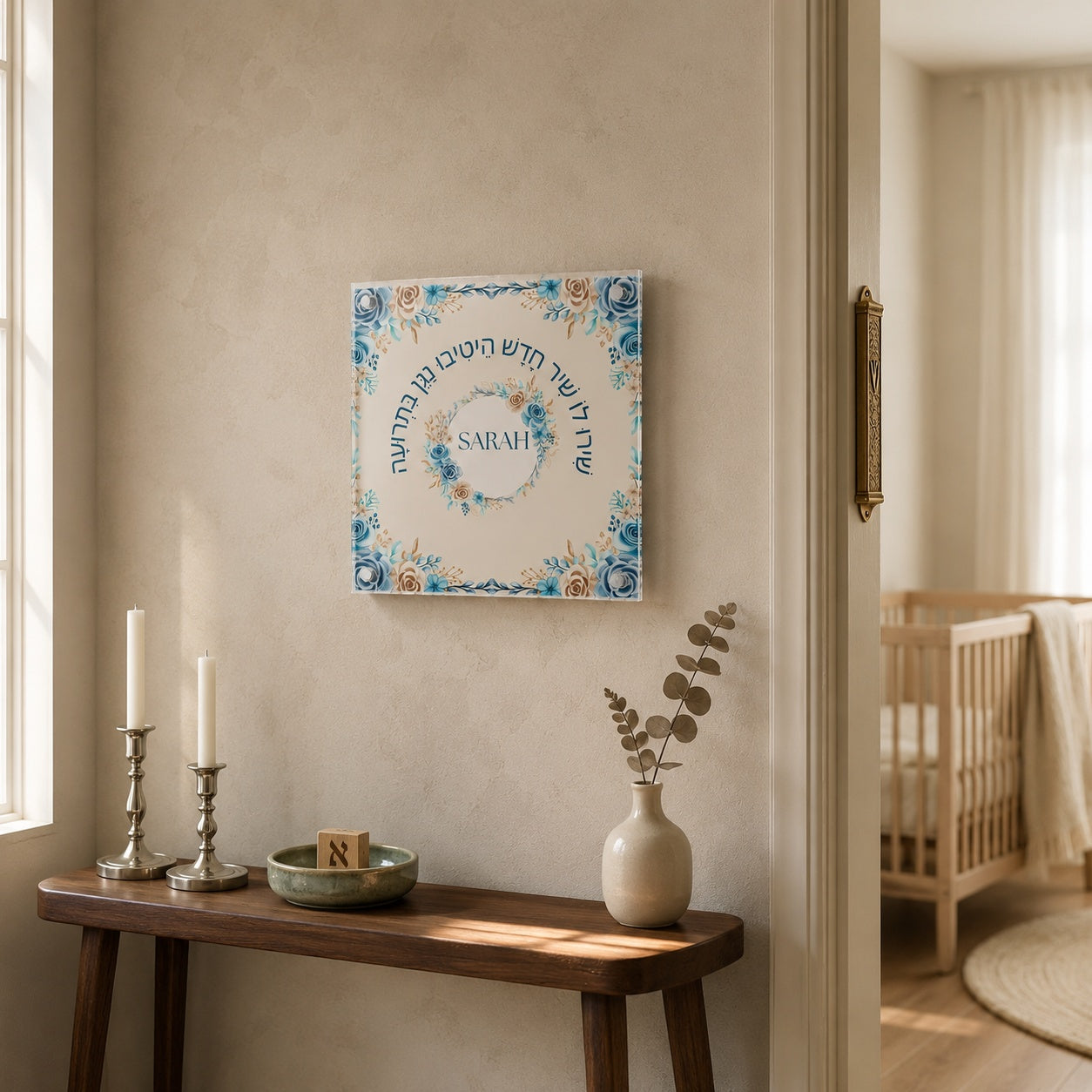 Framed Hebrew name pasuk art hanging above an oak crib in a sunlit cream nursery with sage accents.