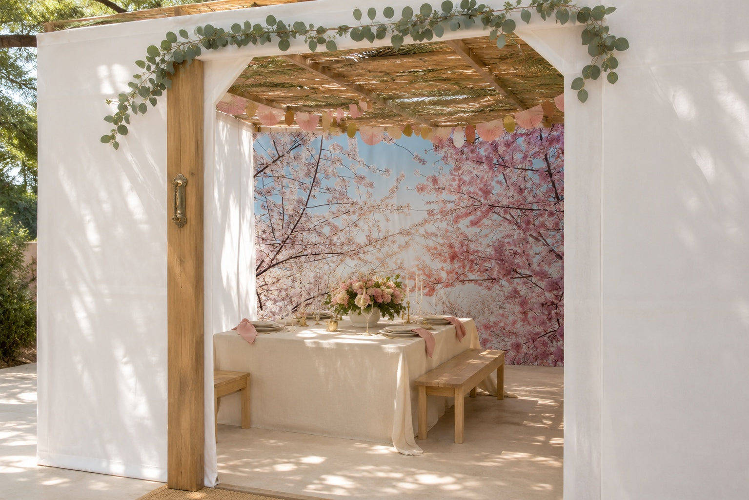 White fabric sukkah with bamboo roof, cherry blossom wall mural behind a set table with wooden benches and pink florals.